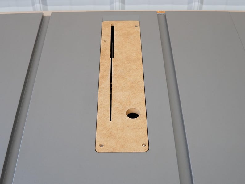 Puede incluir: Primer plano de una mesa de color gris con un recorte rectangular para un elevador de fresadora. El recorte est&aacute; hecho de madera marr&oacute;n claro y tiene un peque&ntilde;o agujero en el centro.