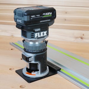 May include: A cordless FLEX 24V router with a black and gray body, mounted on a black base. The router is positioned on a wooden surface next to a silver guide rail with a green stripe. The router has a battery pack on top.