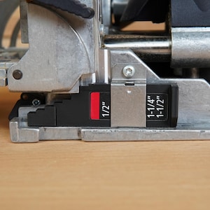 May include: Close-up of a metal woodworking tool with black and red markings. The tool has measurements of 1/2", 1-1/4", and 1-1/2" marked on it. The tool is set on a light brown wooden surface.