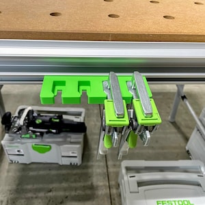 May include: Green and silver tool holder with three clamps hanging from it. The holder is attached to a silver track system.