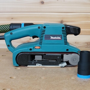 May include: A teal blue belt sander with a black dust port and a black dust collection hose. The sander has a black belt and a silver metal plate. The brand name "Makita" is printed on the sander. A black dust collection adapter is in the foreground.