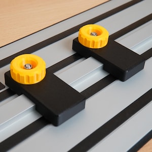 May include: Close-up of a DeWalt track saw guide rail system. The system features a silver rail with black accents, two black brackets, and yellow knobs. The DeWalt logo is visible in the corner. The system is designed for woodworking.