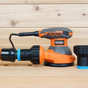 K&ouml;nnte beinhalten: Ein orangefarbener und grauer Ridgid Exzenterschleifer mit einem schwarzen Netzkabel und einem Staubabsaugschlauch. Ein schwarzer Staubabsaugadapter mit blauem Ring ist ebenfalls enthalten. Der Schleifer steht auf einer Holzoberfl&auml;che.