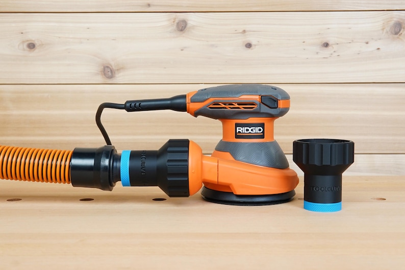 K&ouml;nnte beinhalten: Ein orangefarbener und grauer Ridgid Exzenterschleifer mit einem schwarzen Netzkabel und einem Staubabsaugschlauch. Ein schwarzer Staubabsaugadapter mit blauem Ring ist ebenfalls enthalten. Der Schleifer steht auf einer Holzoberfl&auml;che.