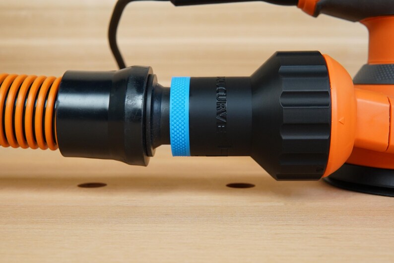 K&ouml;nnte beinhalten: Nahaufnahme eines orange-schwarzen Elektroschleifers mit angeschlossenem Schlauch. Der Schlauch ist orange und gewellt und verbindet sich mit einem schwarzen Adapter mit blauem Ring. Der Schleifer ist orange und schwarz.