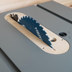 May include: A table saw with a blue circular saw blade. The blade is partially visible through a beige insert in the grey table surface. The word "HER" is visible on the blade. The saw is designed for woodworking.