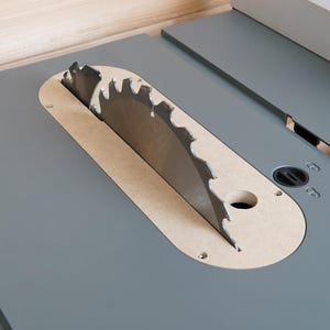 May include: Close-up of a table saw blade. The circular saw blade is silver and has sharp teeth. It is partially visible through a tan-colored insert on a gray table surface. The saw is designed for woodworking.