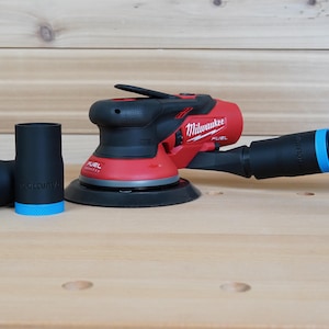 Puede incluir: Una lijadora orbital Milwaukee FUEL roja y negra con una manguera de aspiración negra y dos adaptadores de aspiración negros con detalles azules. La lijadora está sobre una superficie de madera clara.