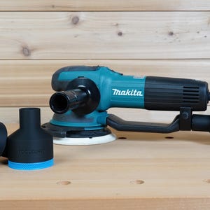 Puede incluir: Una lijadora Makita turquesa y negra con un mango negro y una almohadilla de lijado blanca. Dos accesorios negros de recolección de polvo con detalles azules están junto a la lijadora. Los accesorios tienen el texto "TOOLCRAFT".