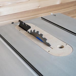 May include: A table saw with a silver blade and a light gray surface. The blade is black and silver with sharp teeth. The saw is set against a light wood background.