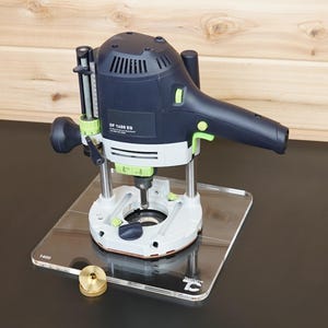 May include: A dark blue and light grey Festool OF 1400 EQ plunge router with green accents, mounted on a clear square acrylic base plate. A small brass-coloured circular guide bushing sits on the base plate near the front left corner.