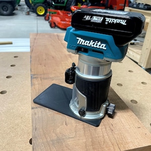 May include: A blue and black Makita cordless router with a black base plate and a 20V Max battery. The router is sitting on a wooden workbench.