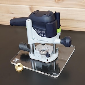 May include: A dark blue and light grey power router mounted on a clear acrylic base plate. The tool features a black handle, a green adjustment knob, and a circular opening at the bottom. A small, circular brass-coloured metal insert sits nearby.