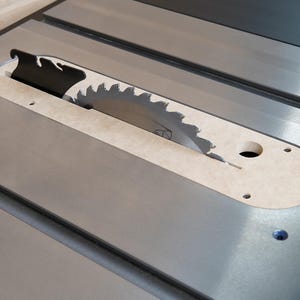 May include: Close-up of a table saw with a circular blade. The blade is partially exposed through a beige insert. The saw's surface is metallic gray, with a black blade guard. The saw is designed for woodworking.