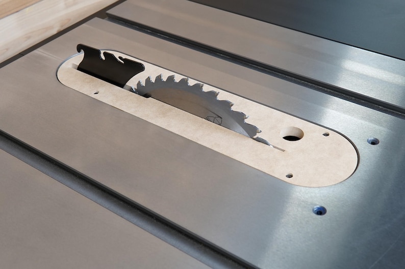 May include: Close-up of a table saw with a circular blade. The blade is partially exposed through a beige insert. The saw's surface is metallic gray, with a black blade guard. The saw is designed for woodworking.