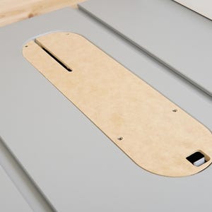 Può includere: Superficie di una sega da tavolo grigio chiaro con un inserto in legno naturale. L'inserto ha una fessura lunga e stretta ed è fissato con piccole viti. La superficie della sega ha un bordo smussato.