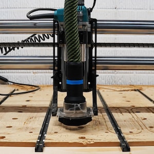 May include: A close-up of a CNC router with a black and blue router bit attached to a green and black vacuum hose. The router is positioned over a piece of wood with a pattern cut into it.