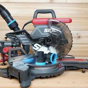 May include: A grey and red SKIL mitre saw with a black handle and a clear blade guard. The saw has a black hose attached and two black and blue attachments. The saw is on a wooden surface.