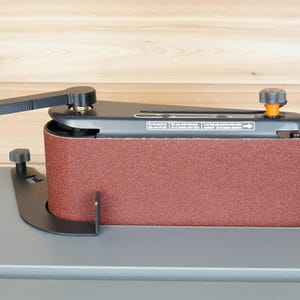 May include: A stationary belt sander with a reddish-brown abrasive belt. The sander has a black frame, a lever, and an adjustment knob with an orange accent. The base is gray, and the background is light brown wood.
