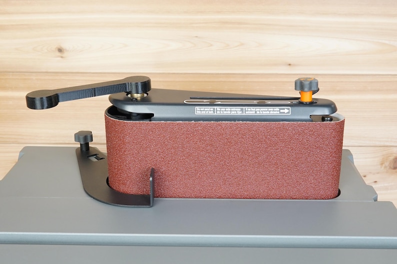 May include: A stationary belt sander with a reddish-brown abrasive belt. The sander has a black frame, a lever, and an adjustment knob with an orange accent. The base is gray, and the background is light brown wood.