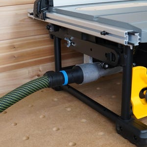 May include: A close-up of a table saw with a black frame, gray table, and yellow accents. A green and black striped hose is connected to the saw. The saw is on a wooden surface.