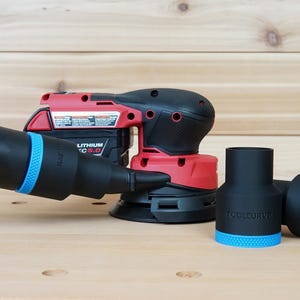 Puede incluir: Una lijadora inalámbrica roja y negra con una manguera negra y adaptadores negros con detalles azules. La lijadora tiene una batería y está sobre una superficie de madera.