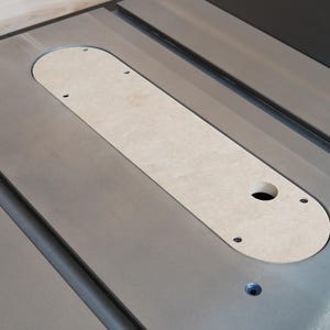 May include: A close-up of a table saw insert plate. The insert is a light tan color, with a rounded rectangular shape. It has a hole and several screw holes. The table saw surface is silver.