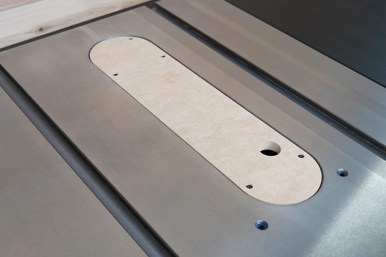 May include: A close-up of a table saw insert plate. The insert is a light tan color, with a rounded rectangular shape. It has a hole and several screw holes. The table saw surface is silver.