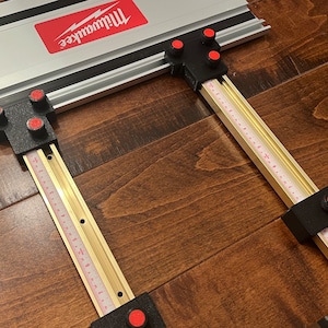 May include: A metal and black Milwaukee track saw guide system. The guide features a silver track with a red Milwaukee logo, black corner brackets, and brass-colored measuring rails with red and white markings. The system is designed for precise cuts.
