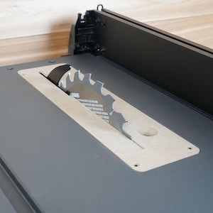 May include: Close-up of a table saw with a silver blade and a gray surface. The blade is circular with sharp teeth. The saw has a black fence and a gray base. The background is a light wood.