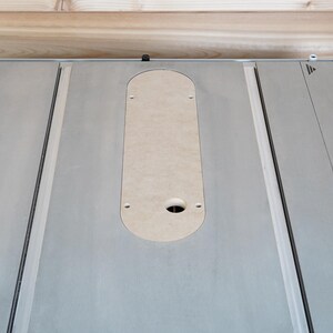 May include: A close-up view of a gray table saw surface with a light-colored, oval-shaped insert. The insert has a hole and several smaller holes. The saw has a black trim and a wooden background.
