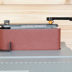 May include: A stationary belt sander with a maroon sanding belt. The sander has a black handle and adjustment knobs. The base is gray, and the background is light brown wood.