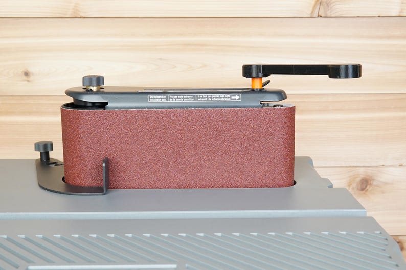 May include: A stationary belt sander with a maroon sanding belt. The sander has a black handle and adjustment knobs. The base is gray, and the background is light brown wood.