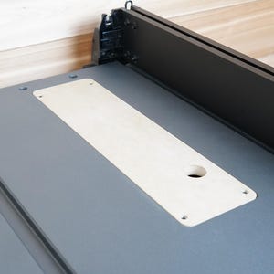 May include: A gray table saw with a black fence and a light-colored insert. The insert has a round hole and is secured with screws. The saw is set against a light wood background.