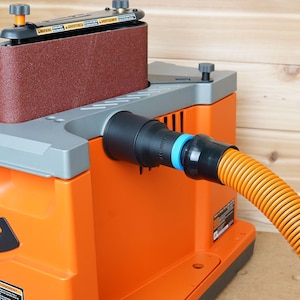 May include: An orange belt sander with a dust collection port and a hose attached. The sander has a brown sanding belt and a grey top.