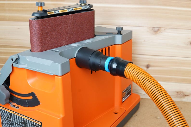 May include: An orange belt sander with a dust collection port and a hose attached. The sander has a brown sanding belt and a grey top.