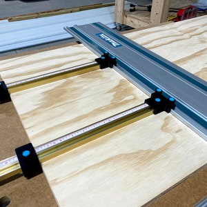 May include: A sheet of plywood is clamped to a workbench using three T-track clamps. The clamps are made of metal and have a black finish with blue accents. The T-track is made of aluminium and has a silver finish with blue accents. The text "T-TRACK" is printed on the T-track.