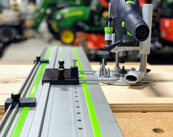 May include: A close-up of a router table with a black and green router attached to a silver track. The track has green lines and a black knob on the left side. The router has a black and silver base with a black and green handle. The text "LM82-1070" is visible on the track.