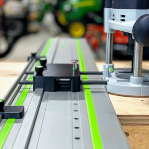 May include: A close-up of a router table with a green and silver track system. The router is attached to the track system and is positioned over a piece of plywood. The router has a black base and a silver top with a green knob.
