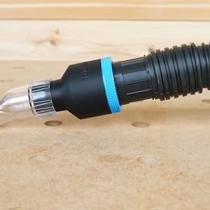 Pode incluir: Uma mangueira de coleta de p&oacute; preta com um adaptador de pl&aacute;stico transparente conectado a uma fresadora. A mangueira tem um anel azul ao redor do adaptador e o texto "TOOL" impresso no lado.
