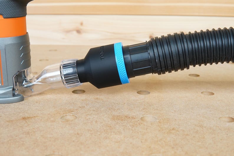 Pode incluir: Uma mangueira de coleta de p&oacute; preta com um adaptador de pl&aacute;stico transparente conectado a uma fresadora. A mangueira tem um anel azul ao redor do adaptador e o texto "TOOL" impresso no lado.