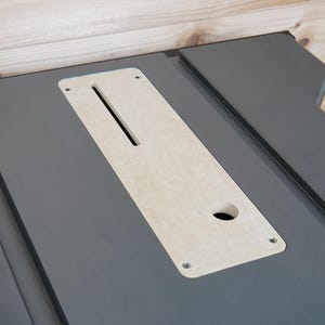 Puede incluir: Primer plano de una sierra de mesa gris con una inserción de color claro. La inserción tiene una ranura larga y estrecha y un agujero circular. La superficie de la sierra es gris mate y la inserción es de color beige claro.