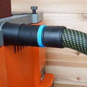 May include: A black and green dust collection hose connected to a sander with a blue and black dust port. The sander has a gray top and an orange base. The text "TOOLCRAFT" is printed on the sander.