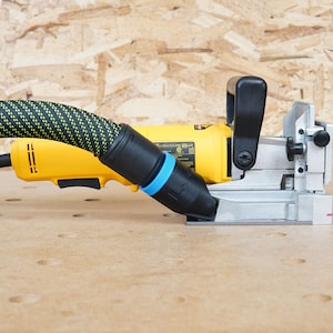 May include: A yellow and black electric router with a dust collection hose attached. The router is sitting on a light brown surface with a few holes in it.
