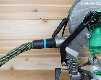 May include: A close-up of a Metabo HPT miter saw. The saw is silver and black with a green blade guard. A black and yellow striped hose is attached to the saw. The background is a light brown wooden surface.