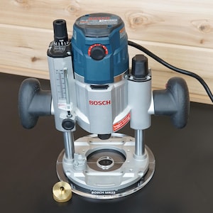May include: A Bosch MR23 plunge router featuring a blue and silver motor housing, two black ergonomic handles, a depth adjustment scale, and a clear circular base plate. A small brass-colored cylindrical component sits next to the tool base.