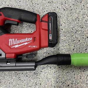 Könnte beinhalten: Eine rote und schwarze Milwaukee FUEL Stichsäge mit einem grünen und schwarzen Staubabsaugaufsatz. Die Stichsäge verfügt über einen Drehregler für die variable Geschwindigkeit und einen Messerfreigabehebel. Der Akku ist mit "M18 RED LITHIUM CP2.0" beschriftet.