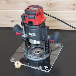 May include: A red and black Skil power router mounted on a clear acrylic base plate. The tool features two ergonomic handles, a digital display on top, and a power cord. A small brass circular component sits on the base plate near the router.