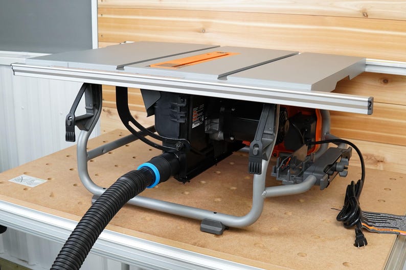 May include: A gray table saw with a black motor and a black dust collection hose. The saw is on a wooden stand with a gray metal frame.