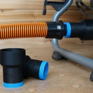 Op de afbeelding: Twee zwarte stofafzuigadapters met blauwe afwerking en de tekst "ToolCury" erop gedrukt. De adapters zijn verbonden met een zwarte en oranje slang.